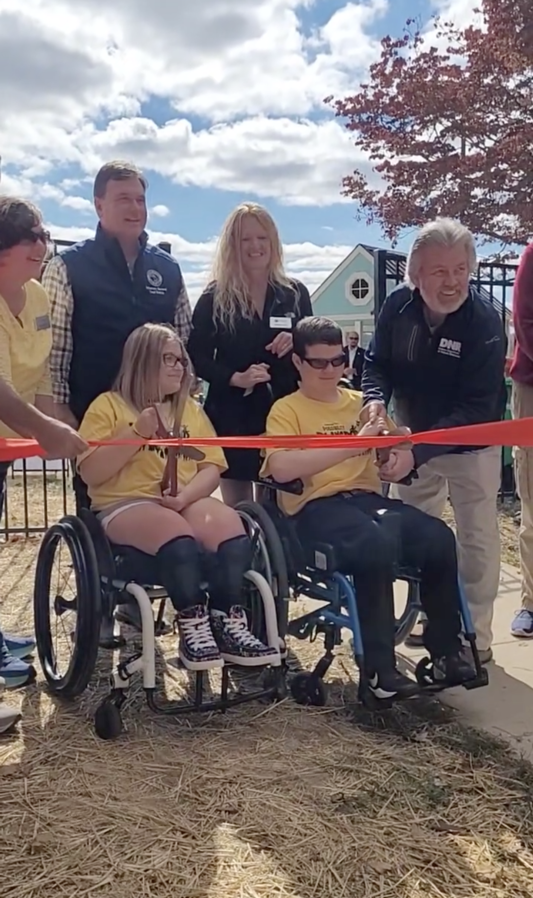 Two children in wheelchairs cut a ceremonial ribbon, with adults standing behind them.